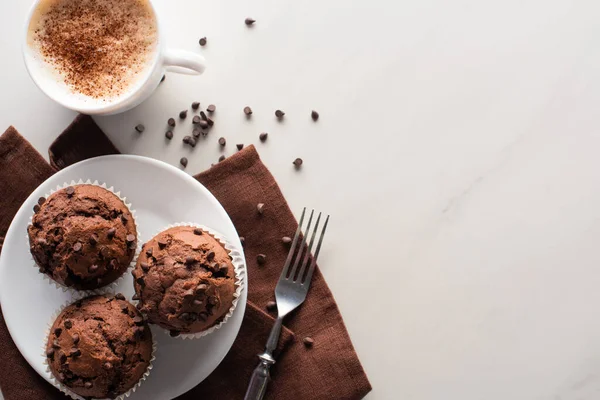 Vista superior de muffins de chocolate fresco na placa branca e guardanapo marrom perto de garfo e cappuccino na superfície de mármore — Fotografia de Stock