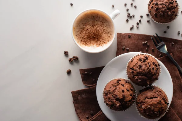 Vista superior de muffins de chocolate fresco na placa branca e guardanapo marrom perto de garfo e cappuccino na superfície de mármore — Fotografia de Stock