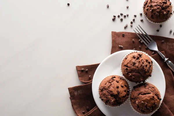 Vista superior de muffins de chocolate fresco na placa branca e guardanapo marrom perto de garfo na superfície de mármore — Fotografia de Stock