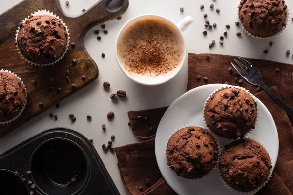 Vista superior de muffins de chocolate fresco na placa de corte, placa branca e guardanapo marrom perto de garfo e cappuccino na superfície de mármore — Fotografia de Stock