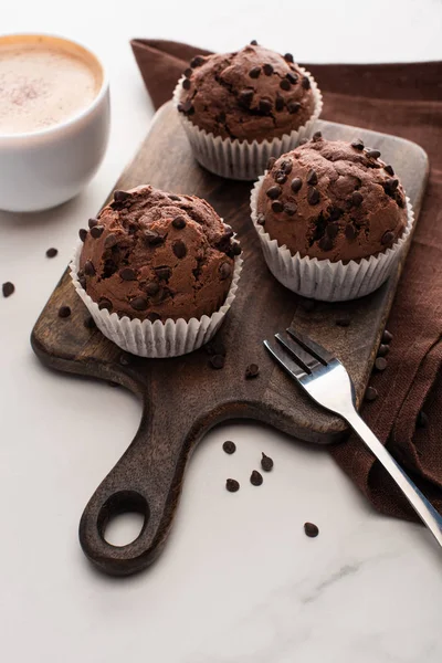 Fresh chocolate muffins on wooden cutting board near napkin, fork and coffee — Stock Photo