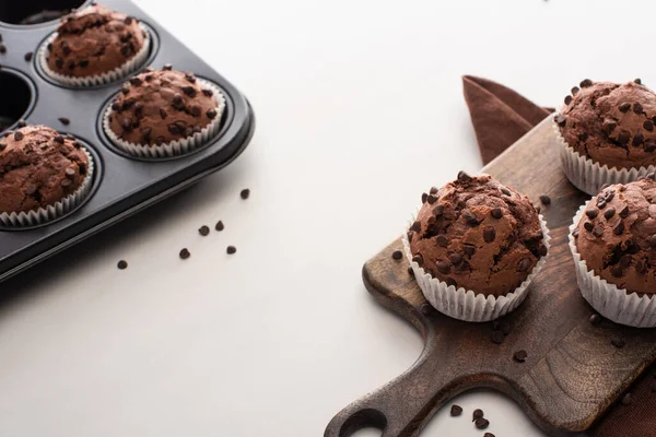 Свежие шоколадные кексы в олове для кексов и на деревянной разделочной доске возле коричневой салфетки — стоковое фото