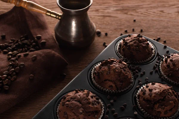 Fresh chocolate muffins in muffin tin on wooden surface near cezve with coffee beans on napkin — Stock Photo