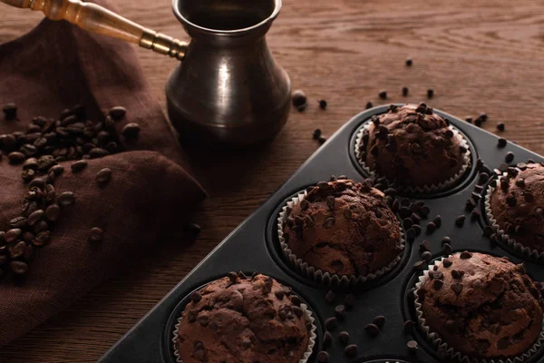 Magdalenas de chocolate fresco en lata de magdalenas en la superficie de madera cerca de cezve con granos de café en la servilleta - foto de stock