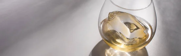 Vetro trasparente con cubetto di ghiaccio e whisky su tavolo bianco con ombra, colpo panoramico — Foto stock