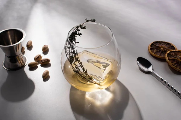 Verre transparent avec herbe, glaçon et whisky sur table blanche avec ombre près de pistaches, cuillère, tranches d'agrumes séchées, gigue — Photo de stock