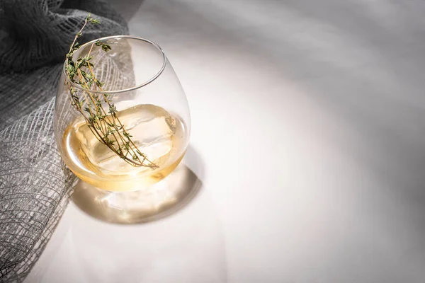 Прозрачное стекло с травой, кубиком льда и виски на белом столе с тенью возле ткани — стоковое фото