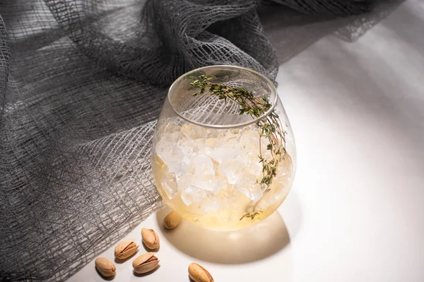 Verre transparent avec herbe, glaçon et whisky sur table blanche avec ombre près du tissu et pistaches — Photo de stock