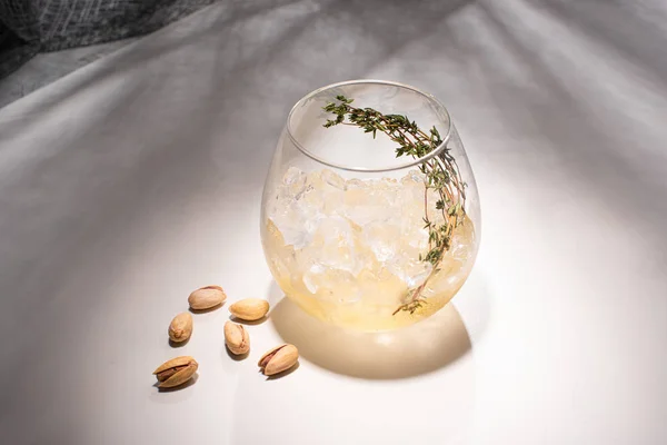Vidrio transparente con hierbas, cubo de hielo y whisky en la mesa blanca con sombra cerca de tela y pistachos - foto de stock