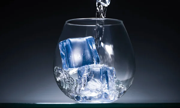 Fresh vodka pouring into transparent glass with ice cubes in dark with blue back light — Stock Photo