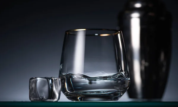 Foyer sélectif de verre transparent avec vodka près de la glace dans l'obscurité avec contre-jour et shaker — Photo de stock
