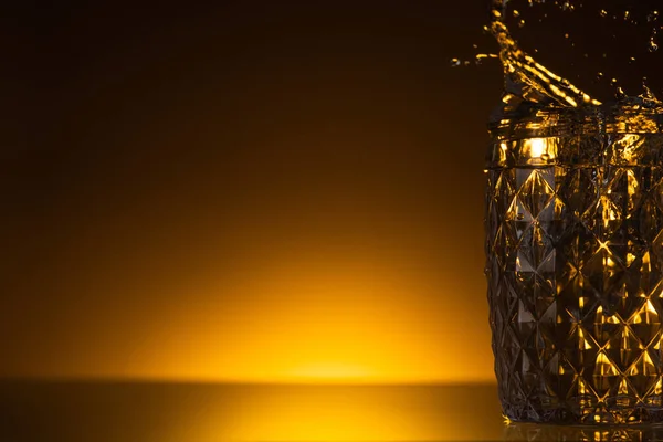 Facettiertes Glas mit spritzendem Wodka im Dunkeln mit warmem Gegenlicht — Stockfoto