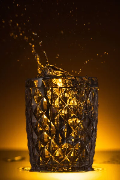 Faceted glass with splashing vodka in dark with warm back light — Stock Photo