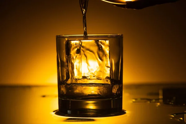 Vodka pouring into transparent glass with ice cube in dark with warm back light — Stock Photo