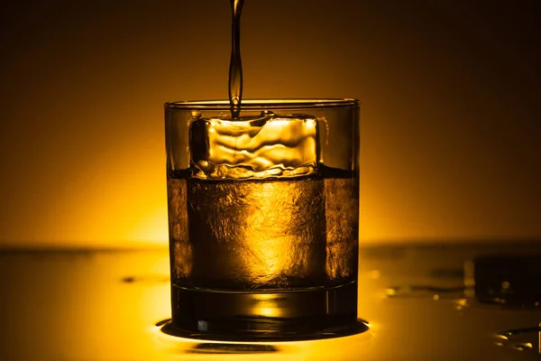 Vodka verter en vidrio transparente con cubo de hielo en la oscuridad con luz de fondo caliente - foto de stock