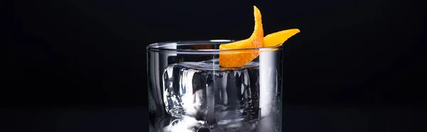Transparent glass with ice cube and vodka garnished with orange peel isolated on black, panoramic shot — Stock Photo