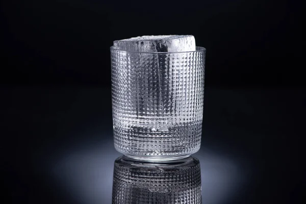 Faceted glass with ice cube on black background — Stock Photo