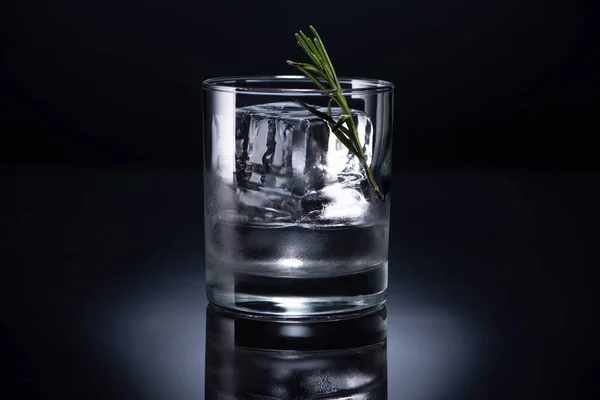 Vidrio transparente con cubo de hielo, romero y vodka sobre fondo negro - foto de stock