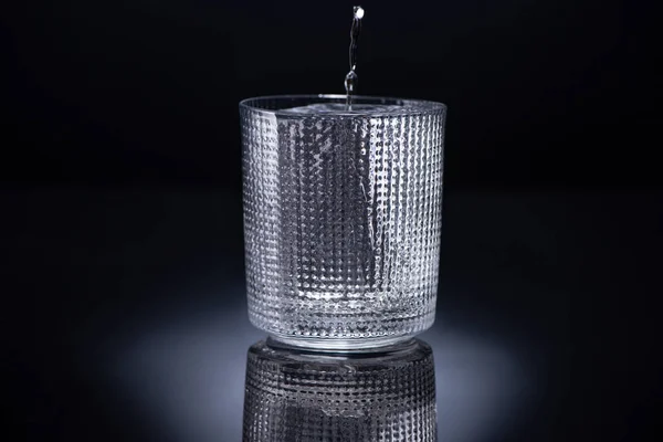 Faceted glass with ice and pouring vodka on black background — Stock Photo