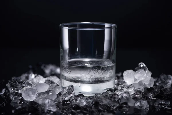 Прозрачное стекло с водкой возле разбитого льда на черном фоне — стоковое фото