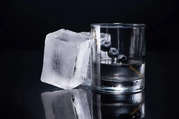 Transparent glass with vodka and blueberries near ice cubes on black background — Stock Photo