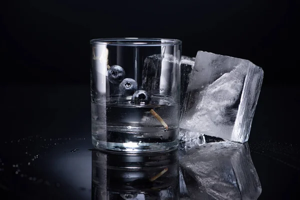 Прозрачное стекло с водкой и черникой возле кубиков льда на черном фоне — стоковое фото