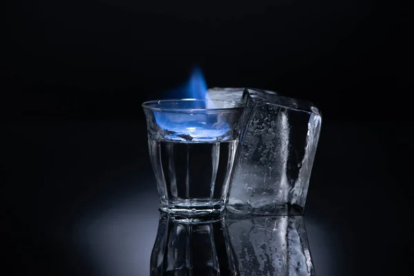 Прозрачное стекло с горящей жидкостью возле льда на черном фоне — стоковое фото