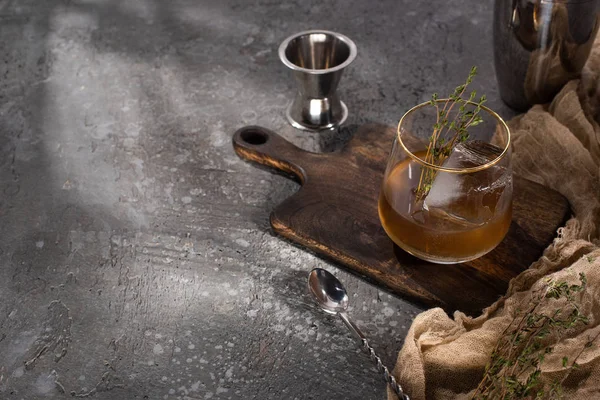 Vidrio transparente con hierba, cubo de hielo y whisky en la superficie de hormigón con equipo de bar - foto de stock