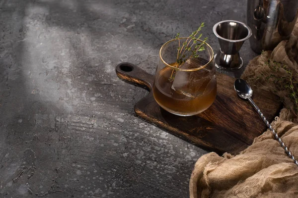 Vidrio transparente con hierba, cubo de hielo y whisky en la superficie de hormigón con equipo de bar - foto de stock