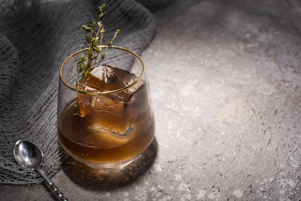 Transparent glass with herb, ice cube and whiskey on concrete surface with mesh cloth and spoon — Stock Photo