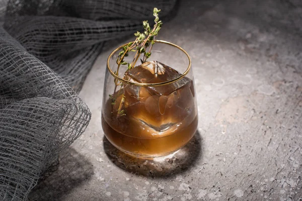 Verre transparent avec herbe, glaçon et whisky sur la surface en béton avec tissu de maille — Photo de stock