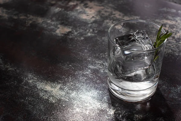 Transparent glass with rosemary, ice cube and vodka on weathered surface — Stock Photo