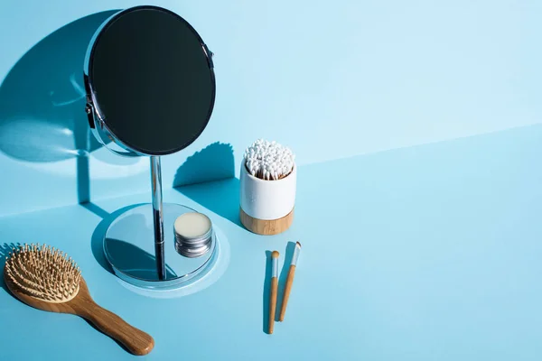 Miroir avec pot de cire à côté de la brosse à cheveux, brosses cosmétiques, porte-brosse à dents avec des bâtons d'oreille sur fond bleu, concept zéro déchet — Photo de stock