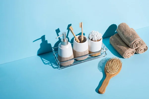 Dispensador, porta cepillos de dientes con cepillos de dientes y orejeras en el estante del baño con toallas, cepillo de pelo sobre fondo azul, concepto de cero residuos - foto de stock