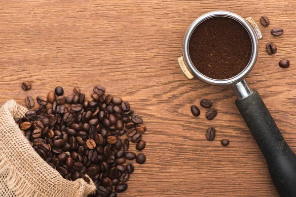 Top view of fresh roasted coffee beans scattered from sack and ground coffee in filter holder on wooden table — Stock Photo