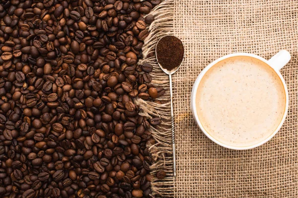 Vista dall'alto di chicchi di caffè tostato fresco e sacco con cappuccino e cucchiaio con caffè macinato — Foto stock