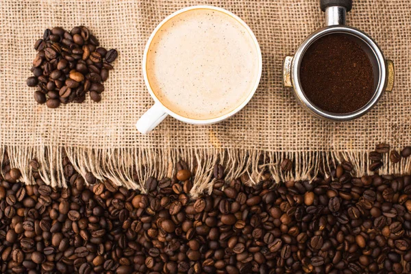 Blick von oben auf frisch geröstete Kaffeebohnen und Sacktuch mit Cappuccino und Filterhalter mit gemahlenem Kaffee — Stockfoto