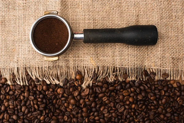 Вид сверху на свежие жареные кофейные зерна и мешковину с держателем фильтра с молотым кофе — стоковое фото