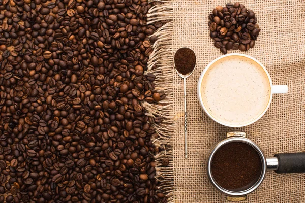 Blick von oben auf frisch geröstete Kaffeebohnen und Sacktuch mit Cappuccino, Löffel und Filterhalter mit gemahlenem Kaffee — Stockfoto