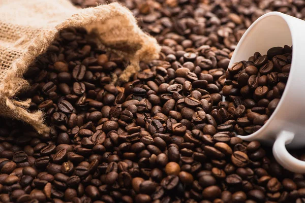 Messa a fuoco selettiva di chicchi di caffè tostato fresco e tazza bianca vicino al sacco — Foto stock