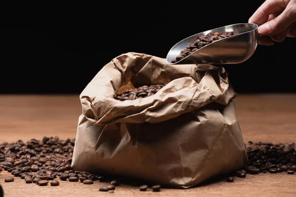 Обрезанный вид человека, держащего лопатку со свежими жареными кофейными зёрнами рядом с бумажным пакетом на деревянном столе, изолированном на черном — стоковое фото