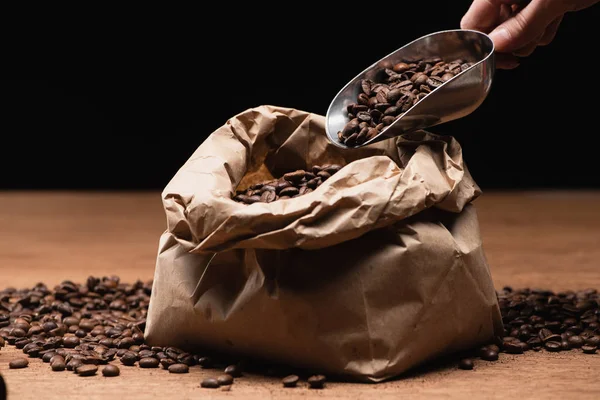 Обрізаний вид людини, що тримає шпатель зі свіжими смаженими кавовими зернами біля паперового мішка на дерев'яному столі ізольовано на чорному — стокове фото