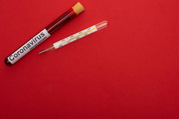 Vista superior del tubo de ensayo con muestra de sangre y letras de coronavirus y termómetro sobre fondo rojo - foto de stock