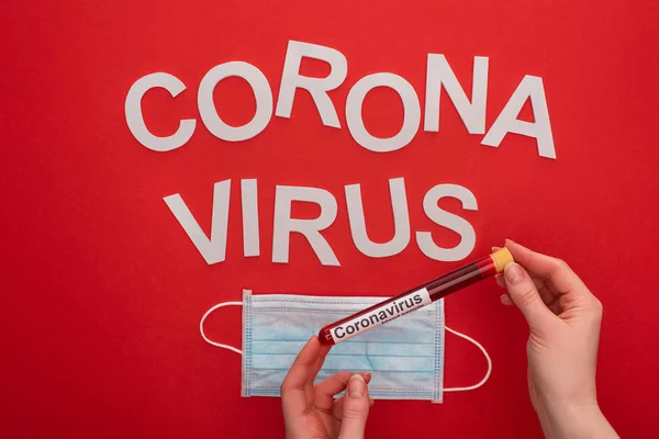 Top view of woman holding test tube with blood sample near medical mask and coronavirus lettering isolated on red — Stock Photo