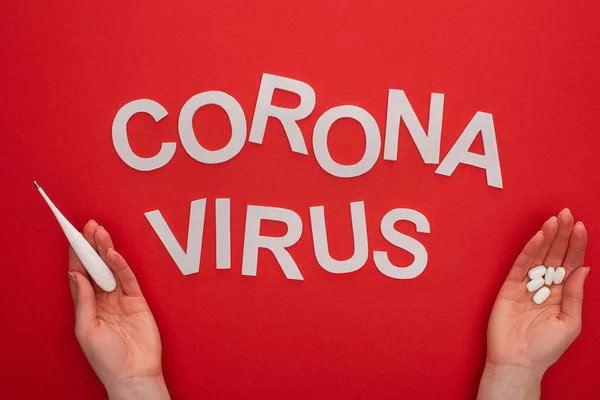 Top view of woman holding thermometer and pills near coronavirus lettering on red background — Stock Photo