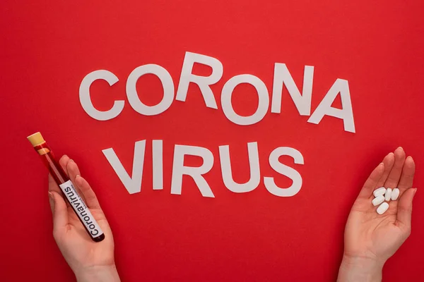 Vista dall'alto della donna che tiene la provetta con campione di sangue e pillole vicino al coronavirus lettering su sfondo rosso — Foto stock