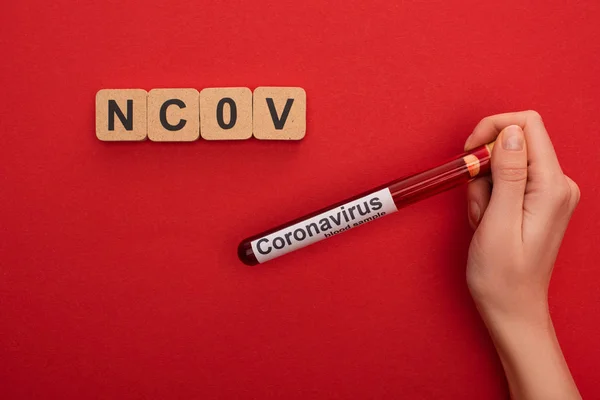 Vista superior de la mujer sosteniendo tubo de ensayo con muestra de sangre y letras de coranavirus cerca de cubos de madera con letras ncov sobre fondo rojo - foto de stock