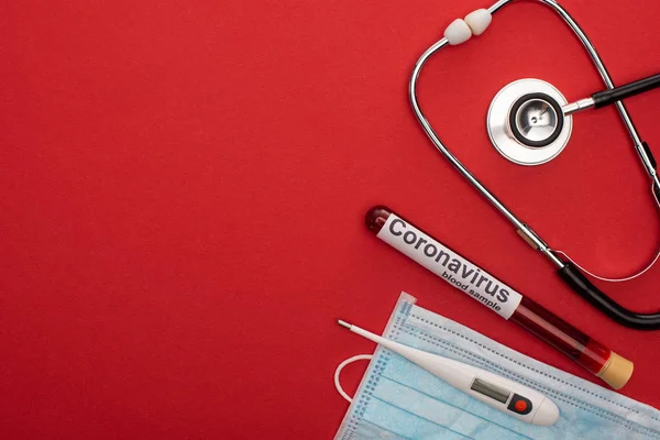 Vista superior del estetoscopio, máscara médica con termómetro y letras de coronavirus en el tubo de ensayo con sangre sobre fondo rojo - foto de stock