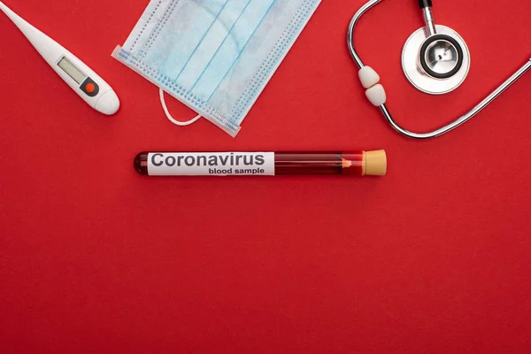 Vista superior de la máscara médica con termómetro, estetoscopio y letras de coronavirus en el tubo de ensayo con sangre sobre fondo rojo - foto de stock