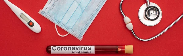 Panoramablick auf medizinische Maske mit Thermometer, Stethoskop und Coronavirus Schriftzug auf Reagenzglas mit Blut auf rotem Hintergrund — Stockfoto